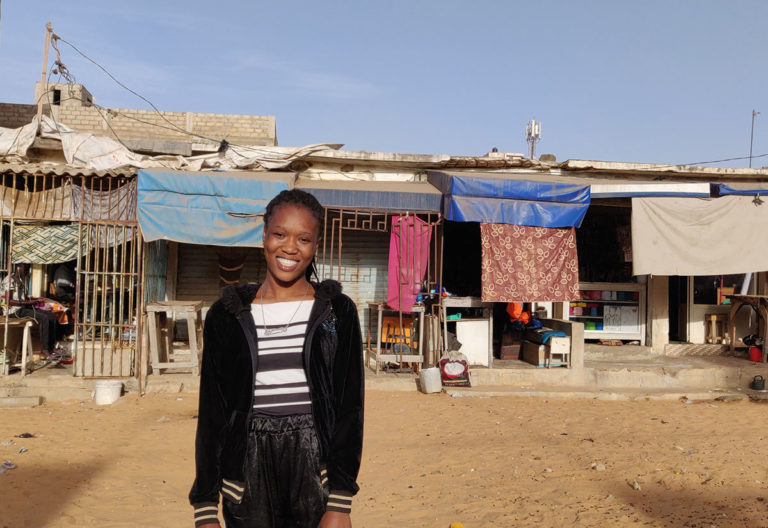 Au Sénégal, une jeune se tient souriante devant des échoppes